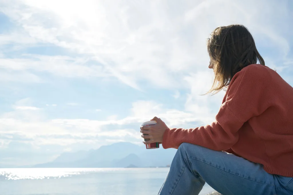 For the Struggling Christian when God Feels Far Away: Woman sitting at the edge of a lake with coffee and thinking