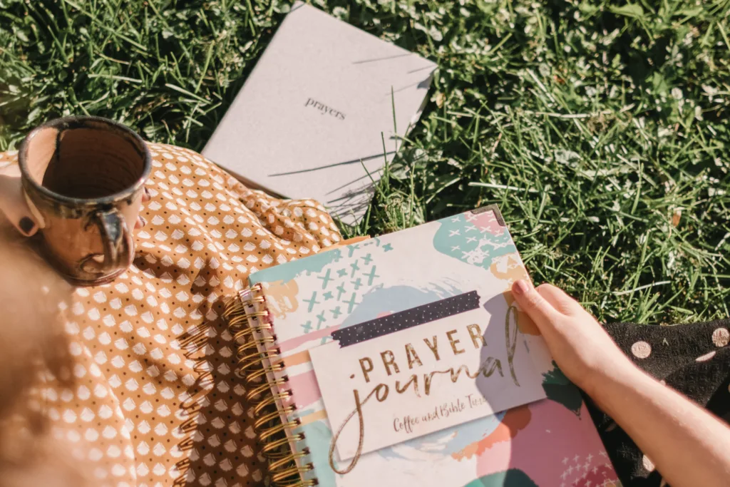 Woman sits in the grass holding her prayer journal ready for prayer journaling.